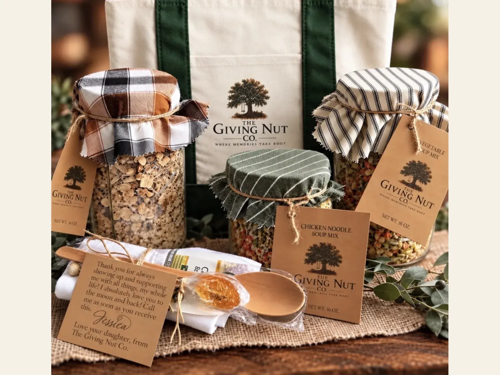 Three Giving Nut Co. jars of soup mix with fabric tops and kraft tags, a canvas tote bag, a wooden spoon and a handwritten gift note, arranged on a burlap runner